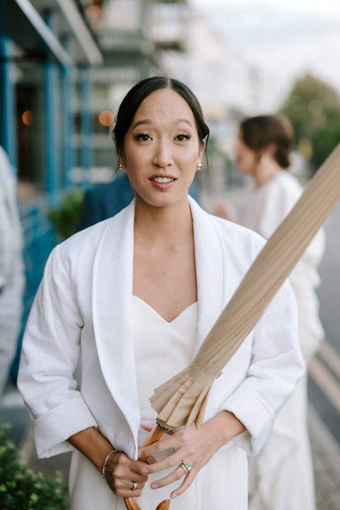 A woman in a white outfit holds a closed beige umbrella, standing outdoors near the Ramsgate Registry Office. Her dark hair is pulled back as she gazes directly into the camera, with blurred buildings and people softly framing her in the background. Image by Pearce Wedding Photography.