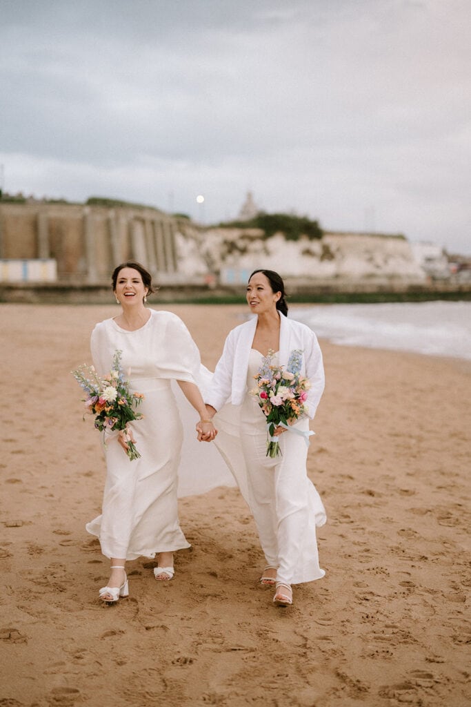 Two women in white attire walk hand in hand on a Ramsgate wedding beach, each holding a colorful bouquet of flowers. The sky is cloudy, and waves gently reach the shore in the background. They are smiling and appear joyful, celebrating their love openly. Image by Pearce Wedding Photography.