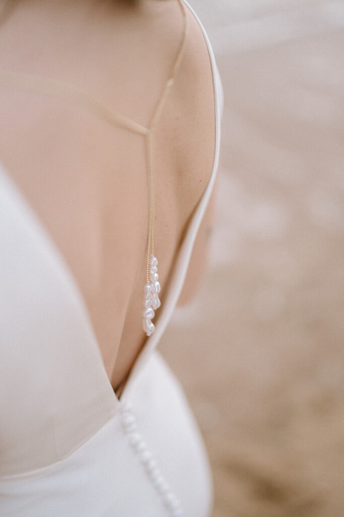 Close-up of a woman in a white dress with a low back, perfect for a Ramsgate wedding, showcasing a delicate strap and line of buttons. The detail includes small pearls cascading from the strap against a softly blurred background. Image by Pearce Wedding Photography.