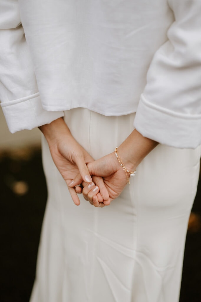 A person stands with their back to the camera at Ramsgate Registry Office, wearing a white long-sleeve top and skirt. Their hands are gently clasped behind their back, adorned with a delicate bracelet, capturing a serene and elegant pose. Image by Pearce Wedding Photography.