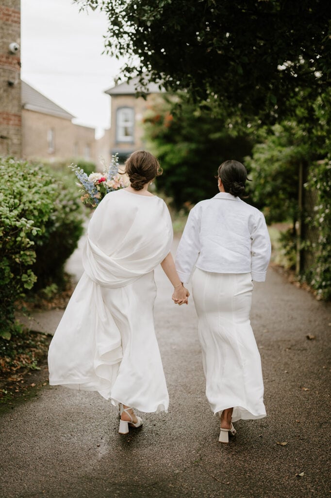 Two people in white outfits hold hands as they stroll down a tree-lined path near the Ramsgate Registry Office. One carries a colorful bouquet, adding charm to the serene scene. It feels like a peaceful residential area, with houses visible in the background. Image by Pearce Wedding Photography.