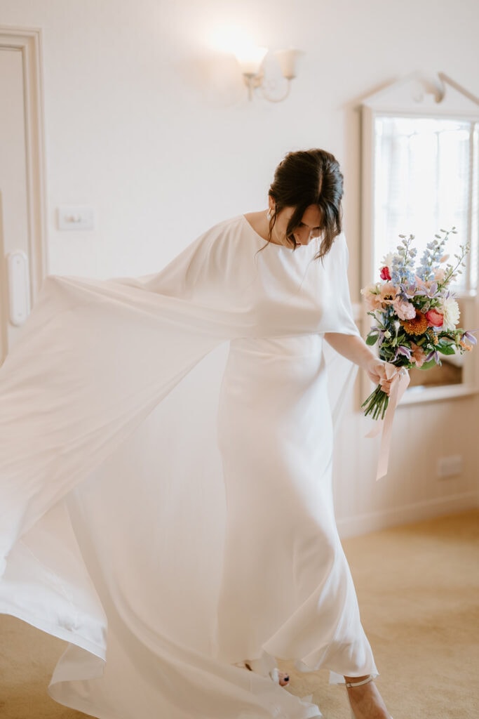A person in a flowing white dress gracefully twirls at the Ramsgate Registry Office, holding a colorful bouquet of flowers. The bright interior space, with soft lighting, a wall sconce, and a mirror, adds a warm, joyful ambiance to the scene. Image by Pearce Wedding Photography.
