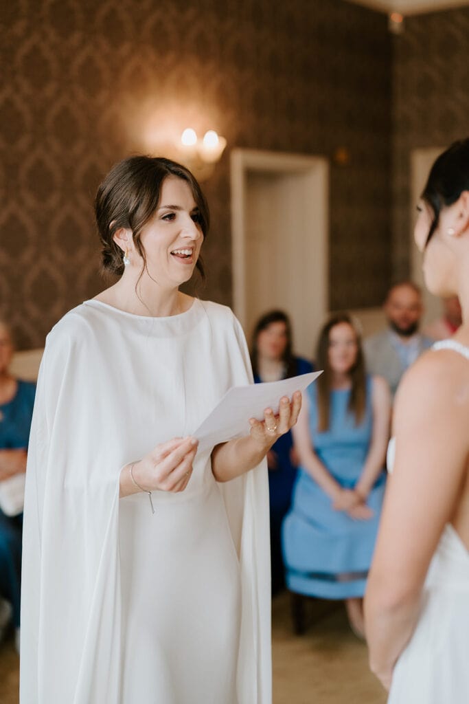 A woman in a white dress is smiling and holding a piece of paper, standing in front of another person at Ramsgate Registry Office. They appear to be in a formal setting with an audience seated in the background. Image by Pearce Wedding Photography.