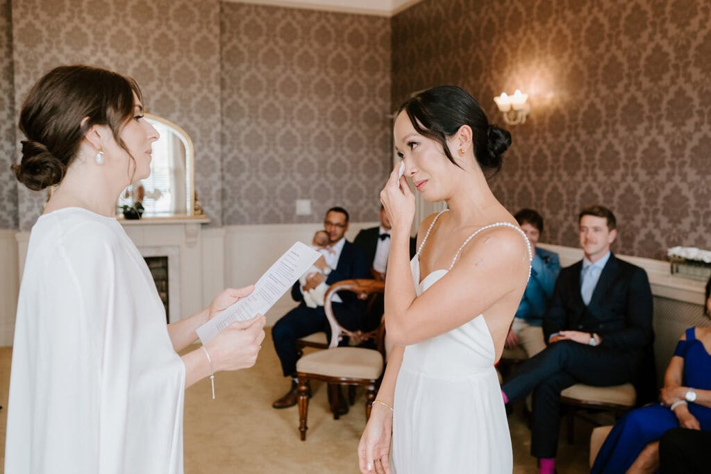 At the Ramsgate registry office, in a room with elegant wallpaper and a soft, warm ambiance, two people stand at the front. One reads vows while the other wipes a tear. Guests seated in the background watch the emotional moment unfold. Image by Pearce Wedding Photography.