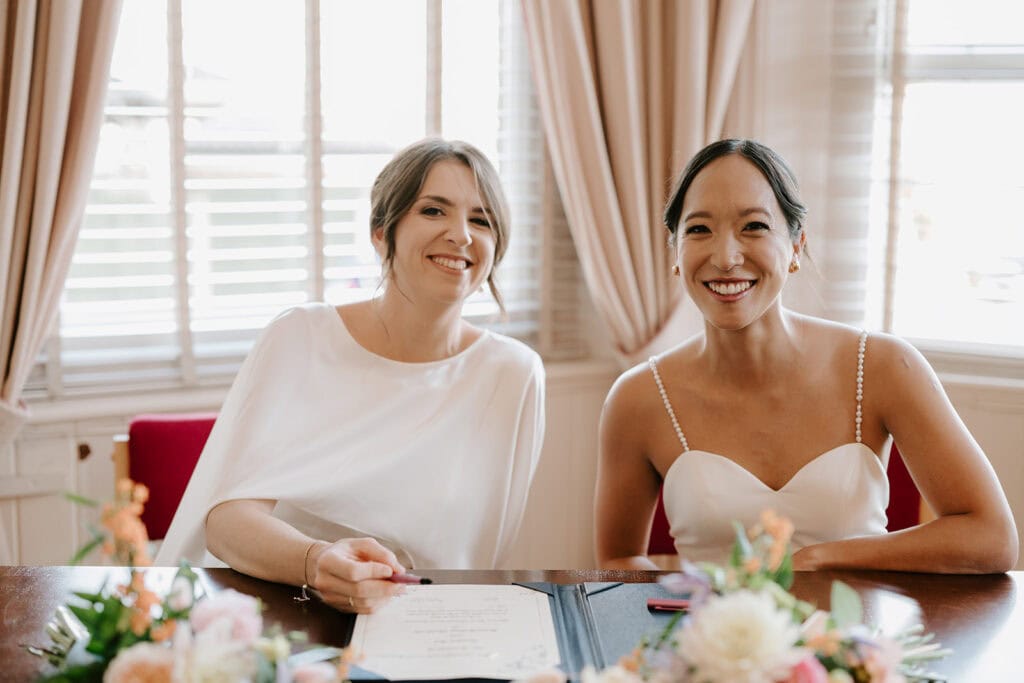 Two women in wedding dresses sit at a table with a bouquet of flowers, smiling at the camera inside the charming Ramsgate Registry Office. The room features light-colored curtains and large windows in the background, adding to the joyful ambiance. Image by Pearce Wedding Photography.