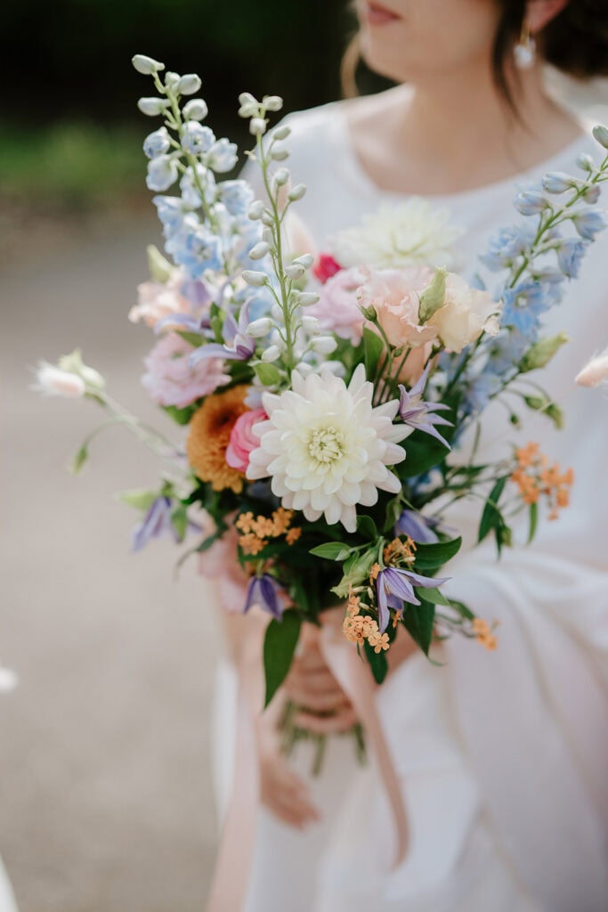 A bride stands in front of Ramsgate Registry Office, holding a colorful bouquet of white dahlias, pink roses, blue delphiniums, and greenery. She wears a white dress against a softly blurred background. Image by Pearce Wedding Photography.