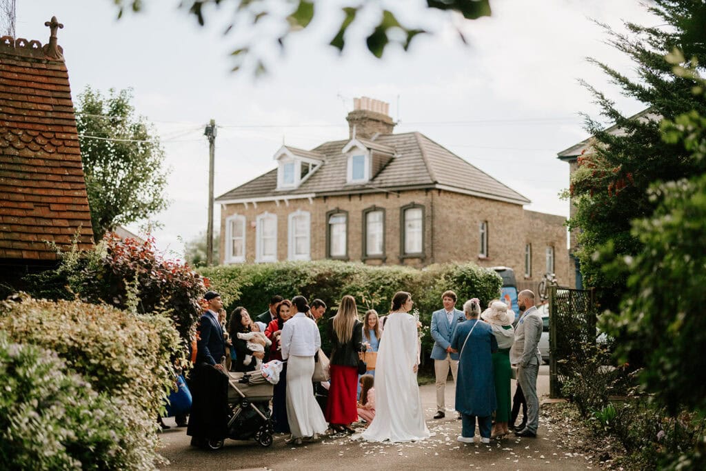 Outside the historic Ramsgate Registry Office, a group of people dressed in formal attire gather near a large brick house. Some are holding babies, and there is a stroller among them. The scene is festive, with petals scattered on the ground and trees lining the path. Image by Pearce Wedding Photography.