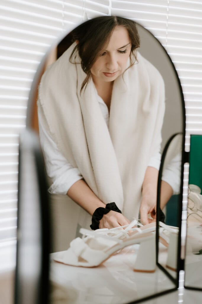 In a softly lit room reminiscent of a Ramsgate registry office, a woman with dark hair wears a white blouse and shawl. She stands in front of a mirror, focused on preparing or organizing items on the table. Image by Pearce Wedding Photography.