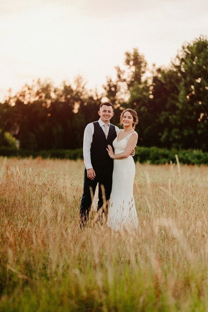 A couple stands in a field at sunset, dressed in formal wedding attire. The woman wears a white gown, and the man wears a vest and tie. Trees are visible in the background.