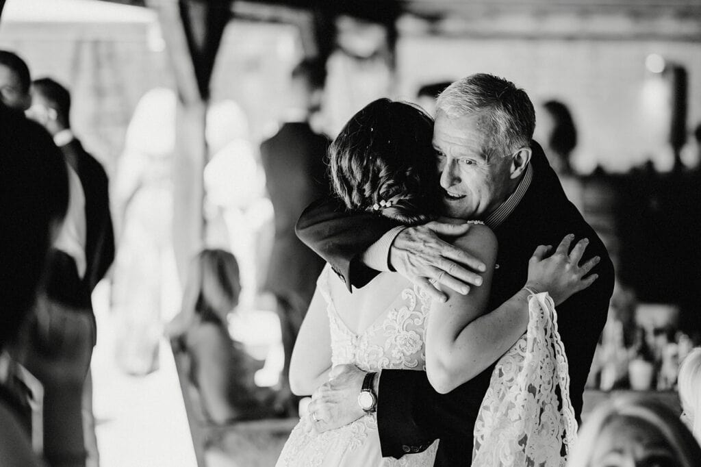 A black and white image of an older man embracing a woman in a lace dress during a gathering.