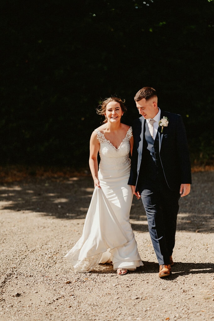 A smiling couple, dressed in a wedding gown and suit, walk outdoors on a sunny day. The woman holds up part of her dress as they stroll together on a gravel path.