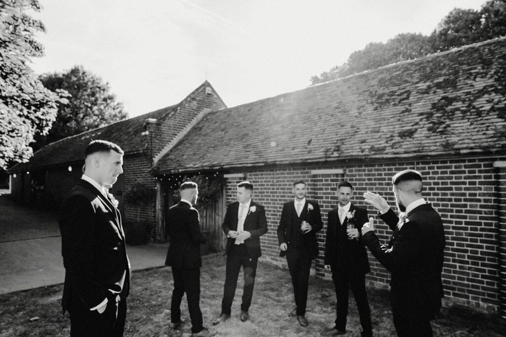 Six men in formal attire stand outside a brick building, engaging in conversation under sunlight.