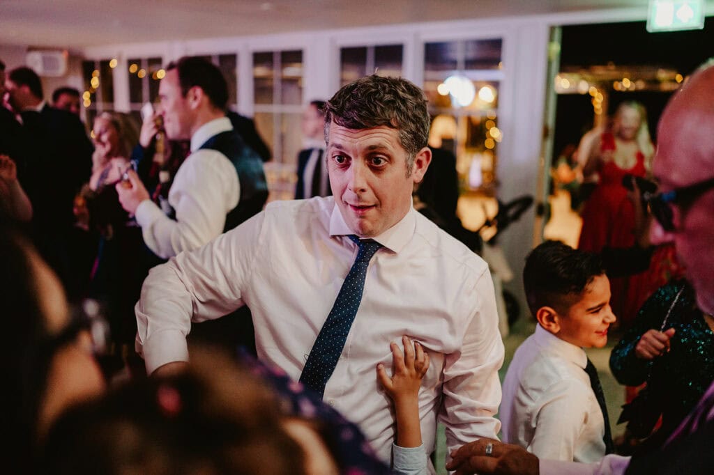 A man in a white shirt and tie is dancing at an indoor event surrounded by other people, including children.