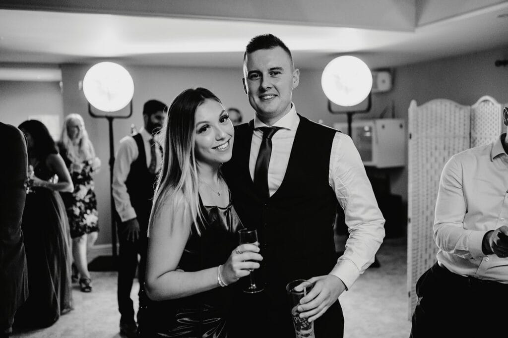 A couple dressed formally, smiling at the camera. The woman holds a drink, and the man has his arm around her. They are in a social setting with other people, lit by round lights in the background.