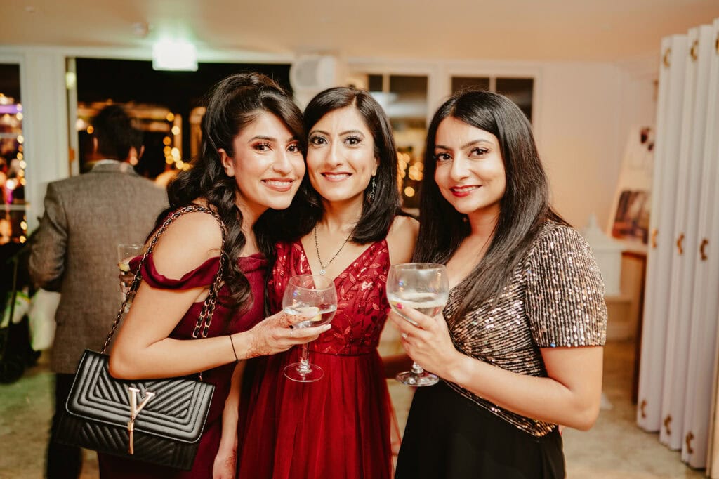 Three women dressed in formal attire hold glasses and smile at the camera in an indoor setting.