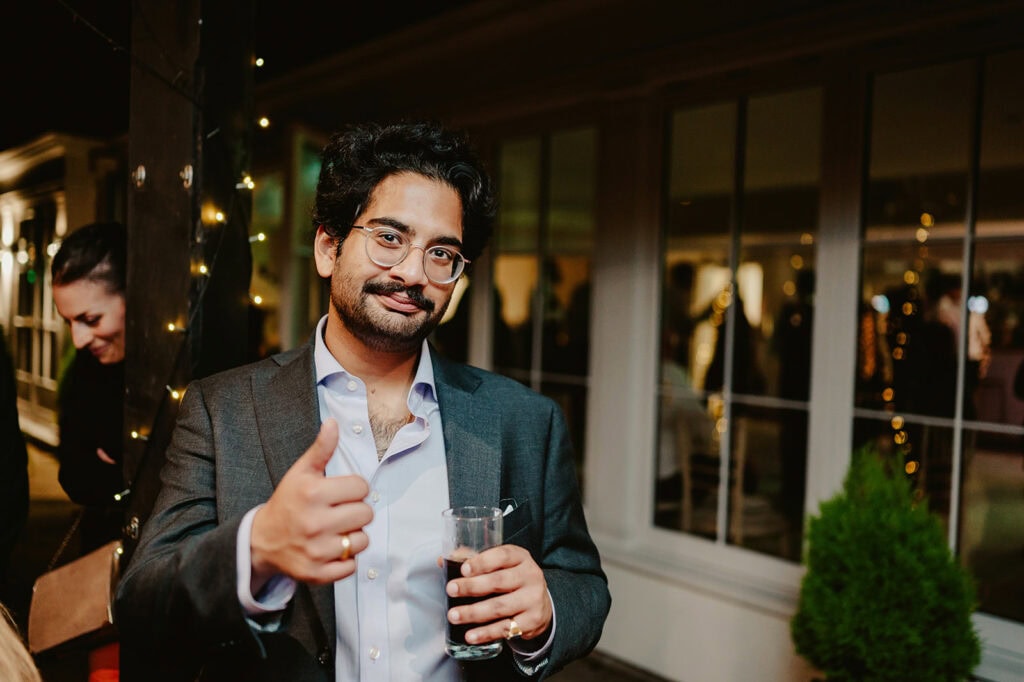 A man in a suit gives a thumbs-up while holding a glass, standing next to a window at a social event.