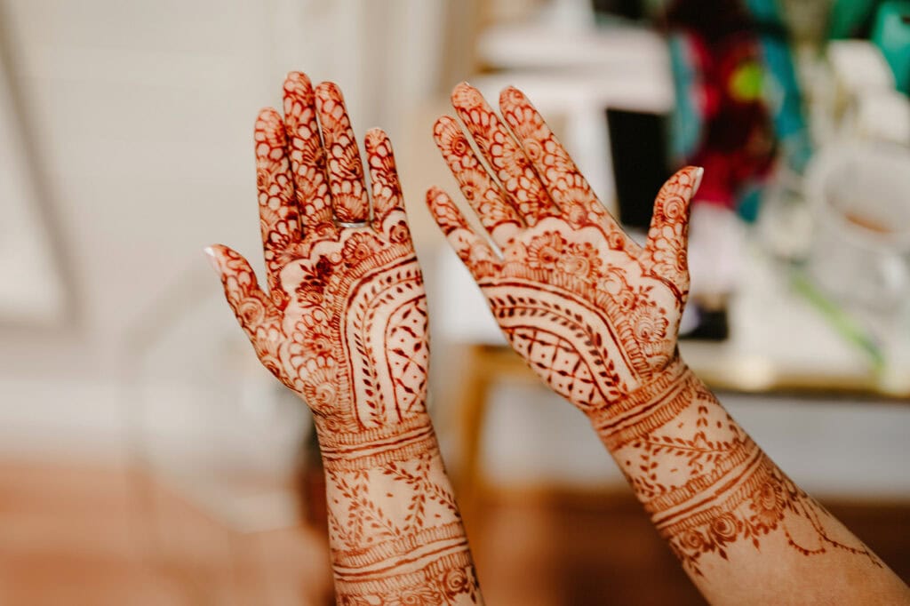 Hands with intricate henna designs on palms and wrists held up with fingers spread apart. Background is blurred.