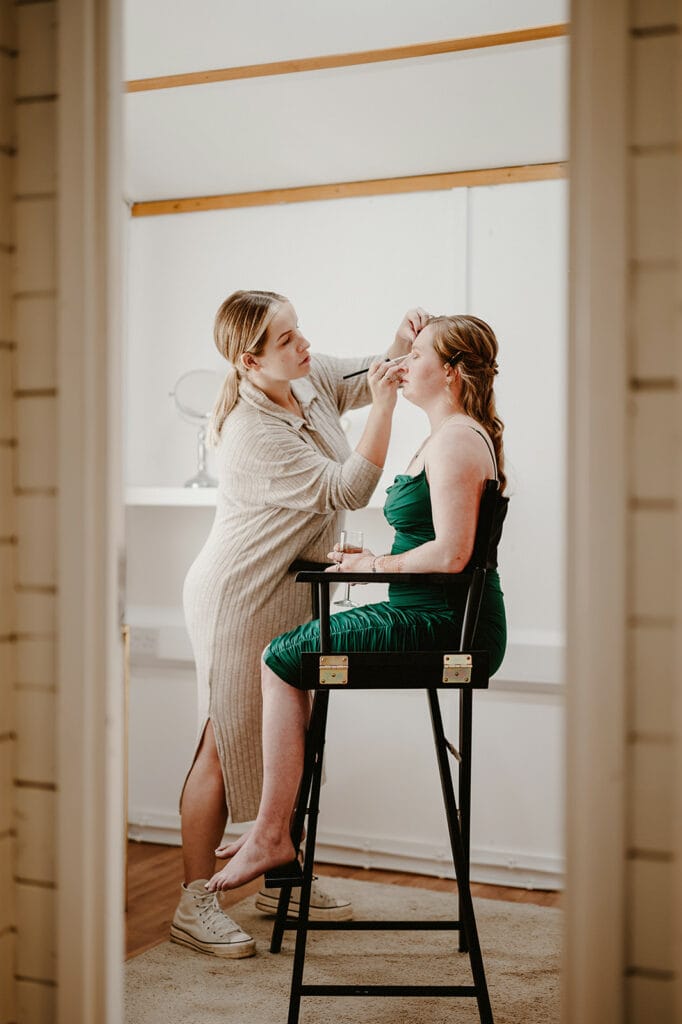 A woman in a beige outfit applies makeup to another woman dressed in a green dress who is seated on a high chair in a well-lit room.