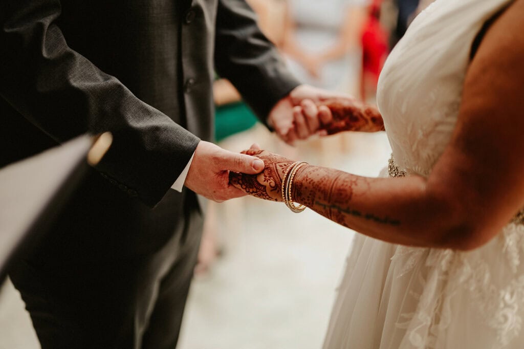 A bride and groom, both dressed formally, are holding hands during their wedding ceremony. The bride has intricate henna designs on her hands.