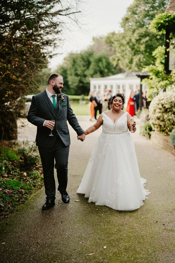 A bride and groom walk hand in hand down a garden path, each holding a glass of wine. The bride wears a white gown, and the groom has a dark suit. Trees and guests appear in the background.