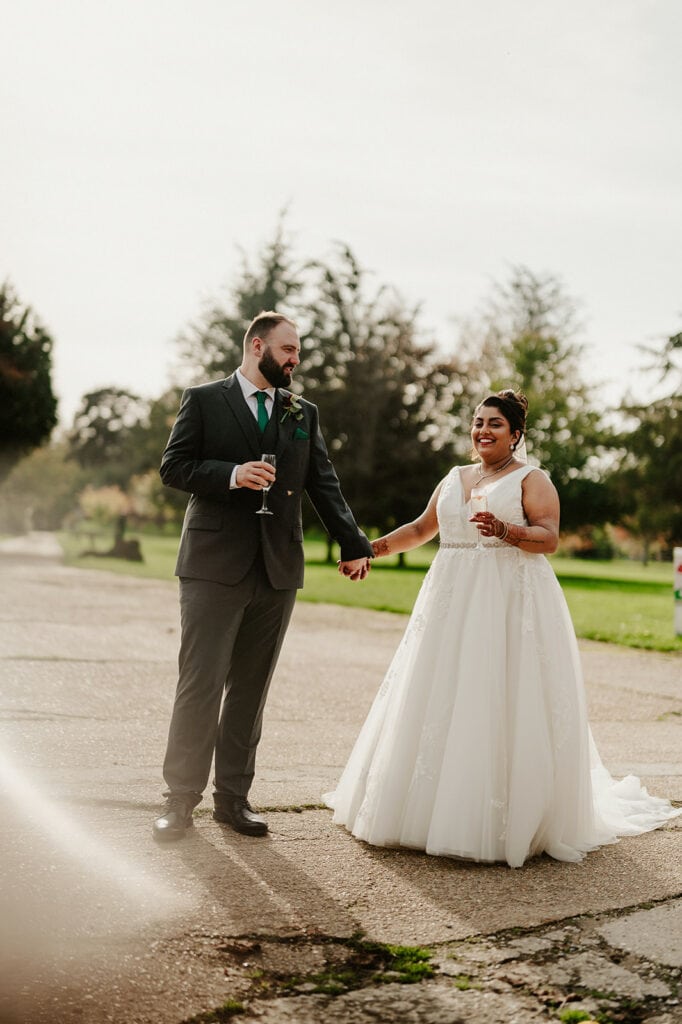 A bride and groom hold hands outdoors. The bride wears a white gown, and the groom is in a dark suit, holding a glass. Trees and a pathway are visible in the background.