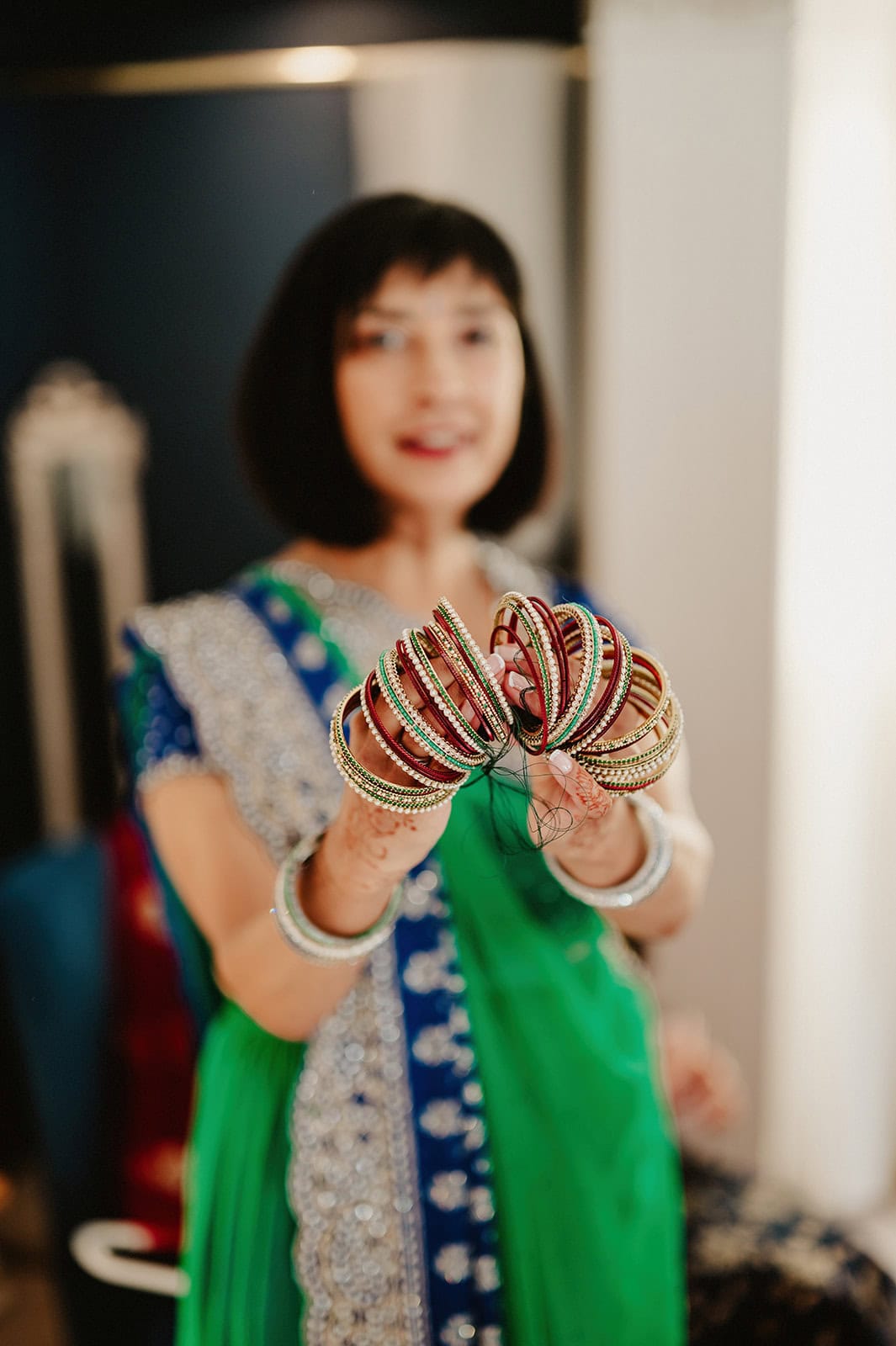 A woman in a green and blue traditional outfit, reminiscent of a scene captured by a Kent wedding photographer, holds out colorful bangles with intricate designs. Her attire features silver accents, and she sports short black hair against a softly blurred background. Image by Pearce Wedding Photography.