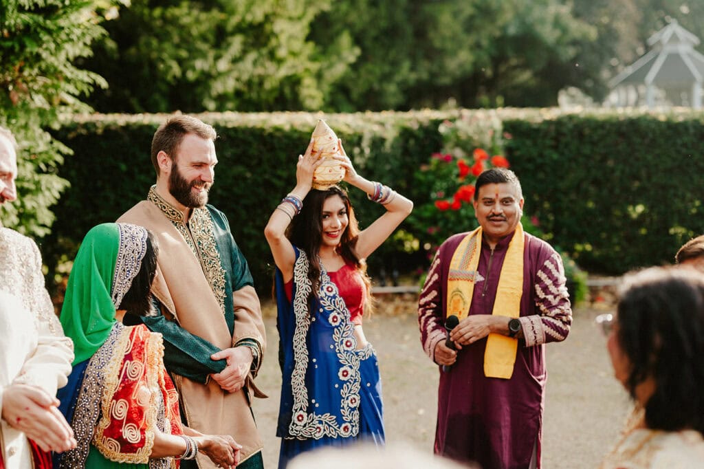 A woman in traditional attire holds an object above her head. She is surrounded by people in colorful clothes, standing outdoors with greenery in the background.