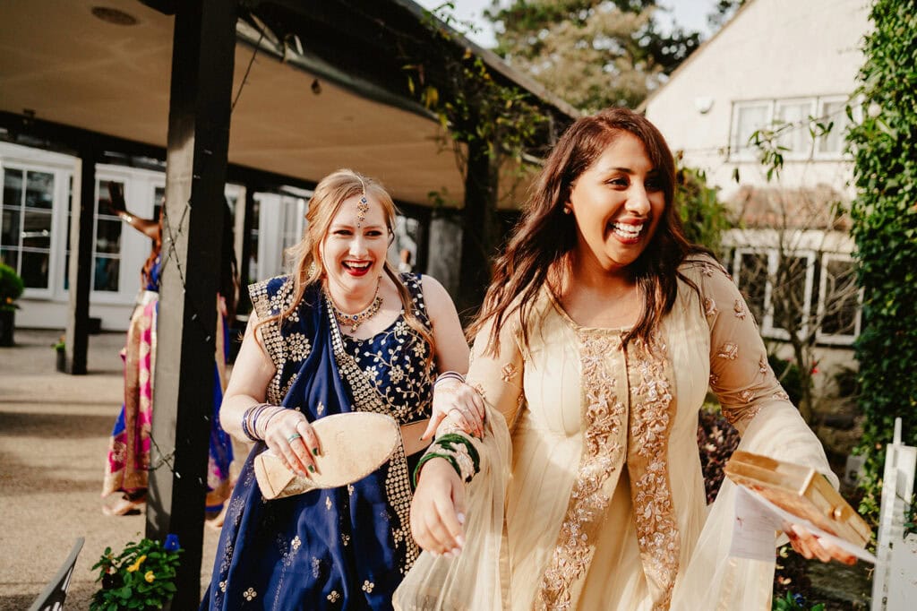 Two women in festive traditional attire laugh and hold hands while walking outdoors during a celebration.