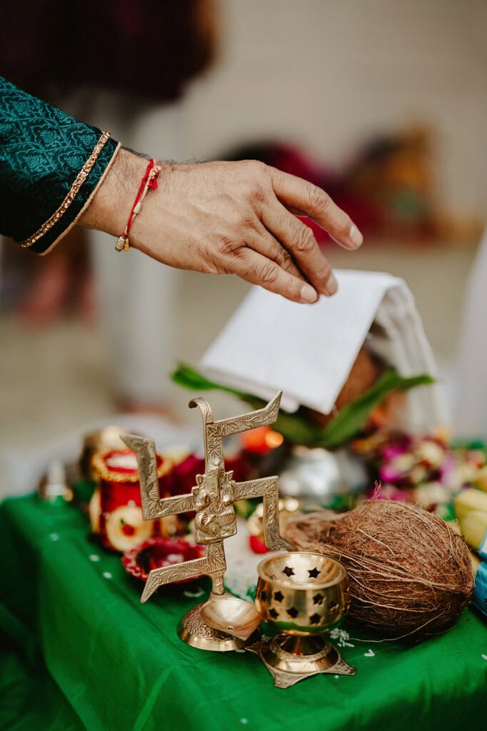 A hand adorned with a bracelet reaches over a table with a green cloth, displaying a coconut, metal ornaments, a paper, and various ceremonial items.