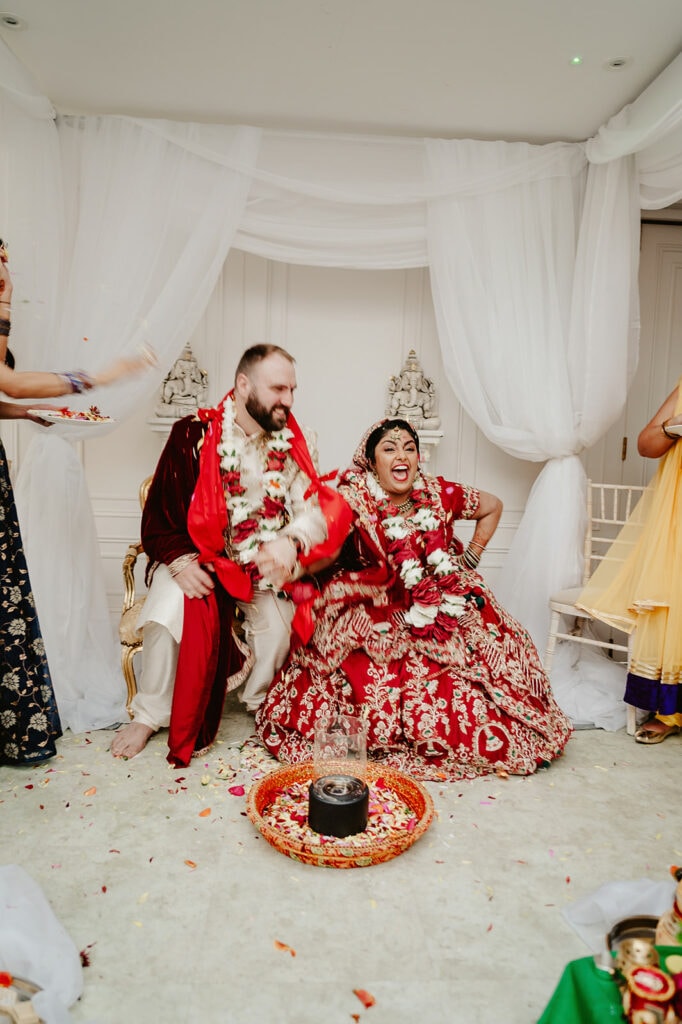 A bride in a red traditional outfit and a groom in ceremonial attire sit joyfully on chairs, surrounded by flower petals and guests, during a wedding ceremony.