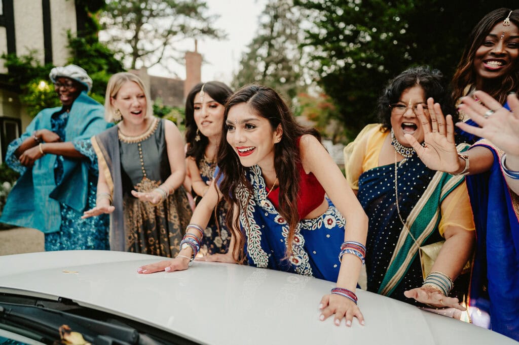 A group of people in festive attire stand around a woman, who is leaning forward with hands on a car, while smiling and engaging with the people around her.