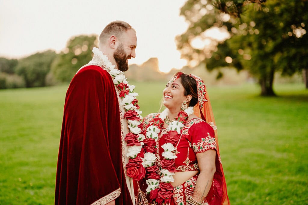 A couple in traditional wedding attire stands outdoors in a grassy area, smiling at each other and wearing floral garlands around their necks.