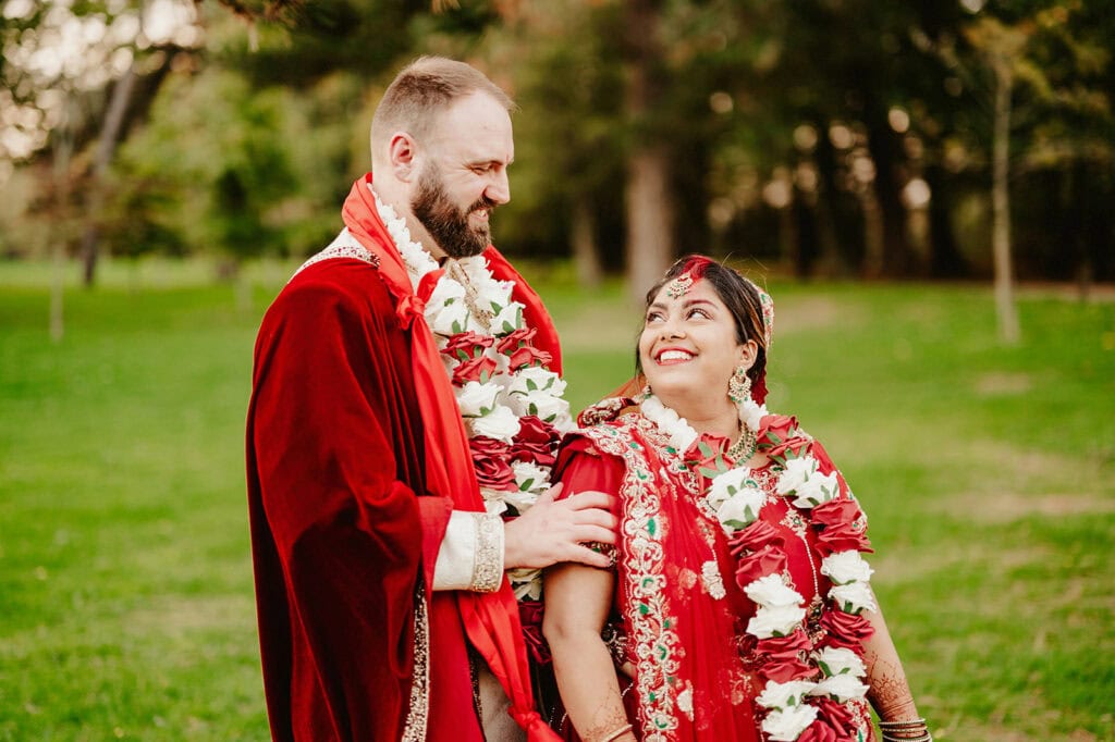 A couple dressed in red traditional attire, adorned with white and red flower garlands, smile at each other while standing outdoors in a green park.