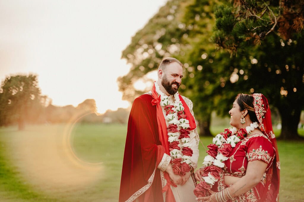 A couple dressed in red and white traditional attire, adorned with floral garlands, stands smiling at each other outdoors near trees during sunset.