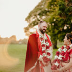 A couple dressed in traditional attire stands in a grassy field at sunset, captured beautifully by a Kent wedding photographer. They wear red garments adorned with floral garlands and jewelry, smiling at each other amidst trees silhouetted against the warm, glowing sky. Image by Pearce Wedding Photography.