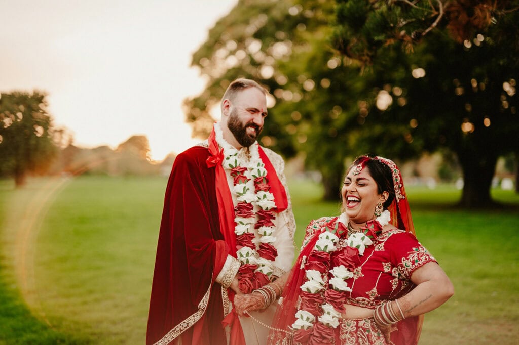 A couple dressed in traditional, vibrant wedding attire stand outdoors, smiling and holding hands with garlands around their necks, against a backdrop of trees and a setting sun.