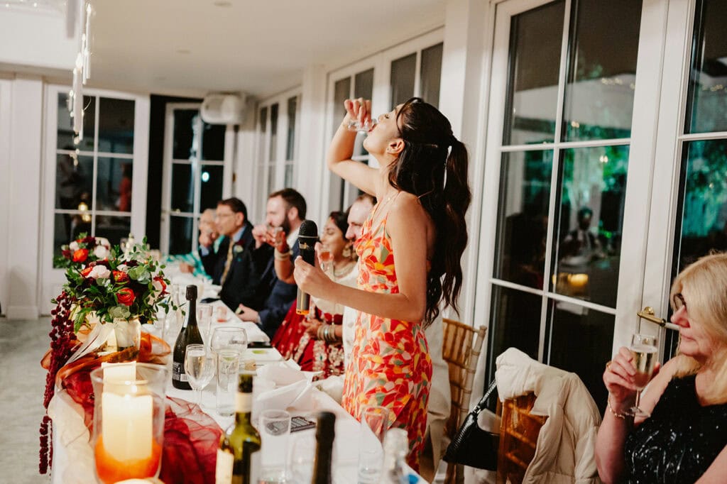 A woman in a floral dress stands at a banquet table, holding a microphone and raising a glass. Other seated guests also raise their glasses. The table is decorated with flowers and lit candles.