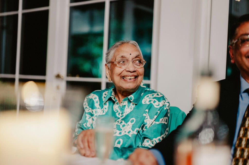 An elderly woman in a green patterned shirt smiles while sitting next to a man in a suit at a table with a candle and glass.