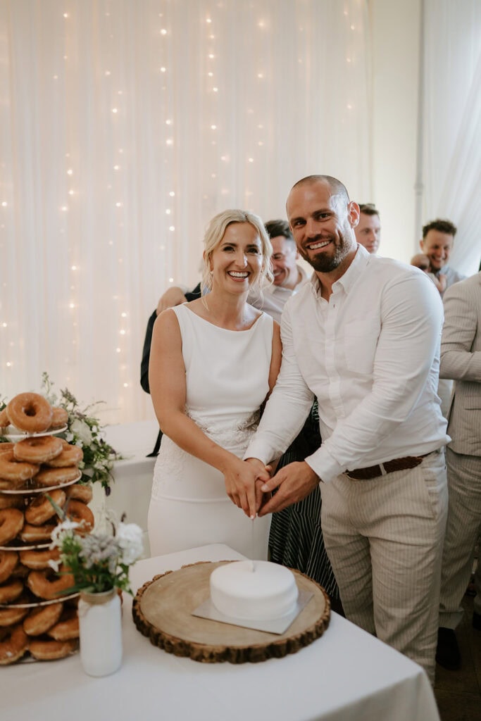 Couple cutting wedding cake with doughnuts nearby.
