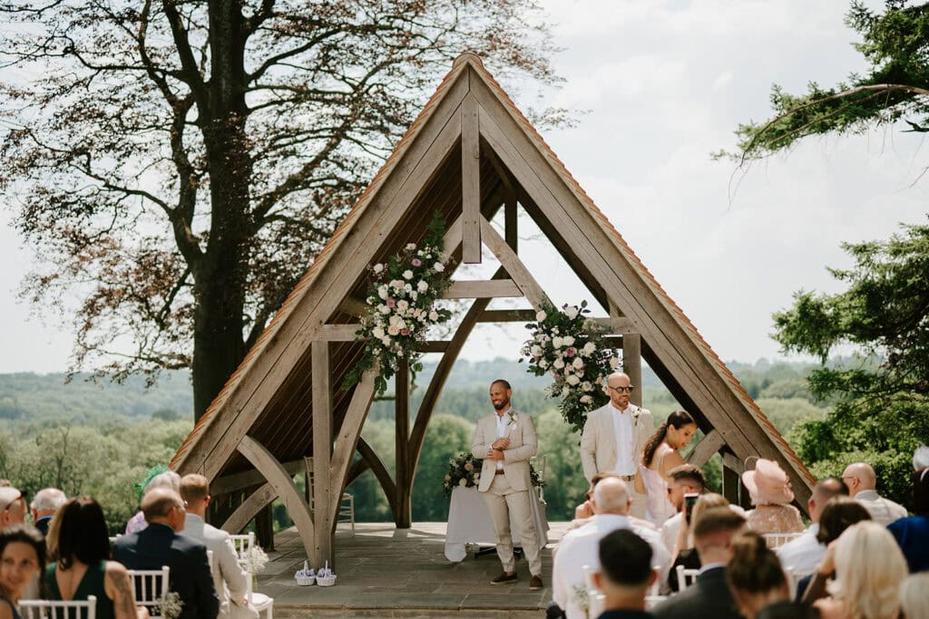 Outdoor wedding ceremony under wooden arch with guests.