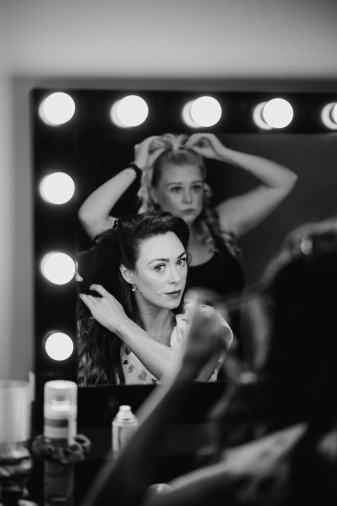 Two women are seen in a dressing room mirror with illuminated bulbs. The woman in front is putting on makeup, while the woman behind is styling her hair, reminiscent of a music photographer's prep space in London. Various beauty products are visible on the counter.