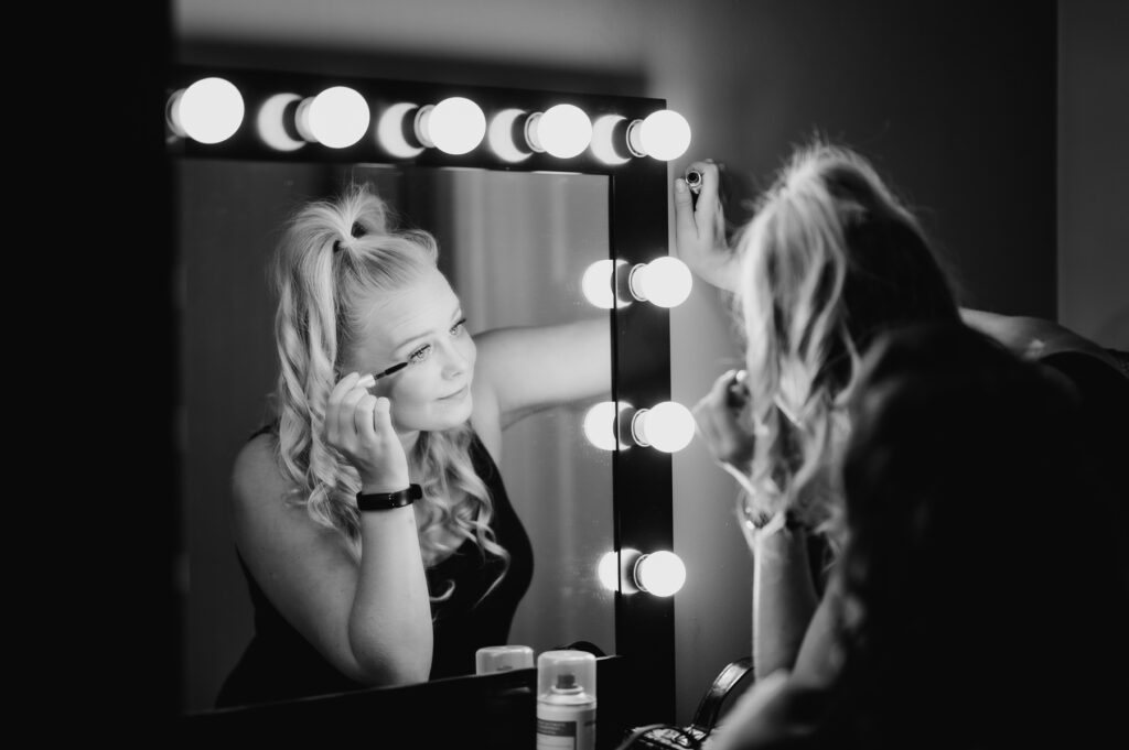 A woman applies mascara in front of a large mirror surrounded by lights. She, a London music photographer with blonde hair tied in a high ponytail, is wearing a dark outfit.