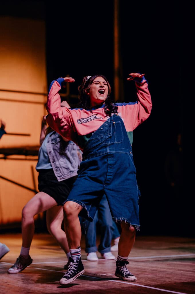 Person in denim overalls and a colorful shirt dancing energetically on a stage as other dancers move in the background; captured by a renowned music photographer from London.