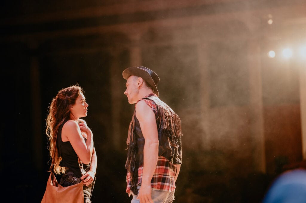 Two people stand facing each other under stage lights. The woman on the left has long hair and wears casual clothing, while the man on the right dons a hat and a fringed vest. Captured by a talented music photographer based in Kent, London, this scene radiates an intriguing harmony of styles.