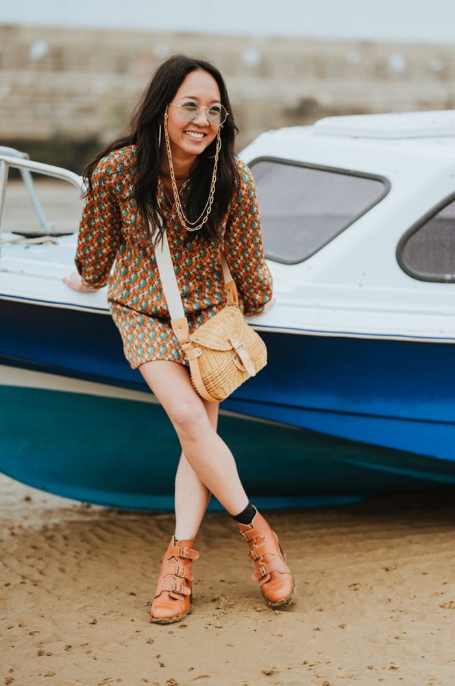 A woman wearing glasses, a patterned dress, and brown boots stands in front of a blue and white boat on a sandy beach. She is smiling and leaning against the boat, holding a woven bag.