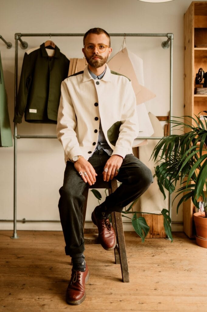 A man with glasses and a beard is seated on a stool in a clothing shop. He is wearing a light-colored jacket, dark pants, and brown shoes. Various garments are displayed in the background.