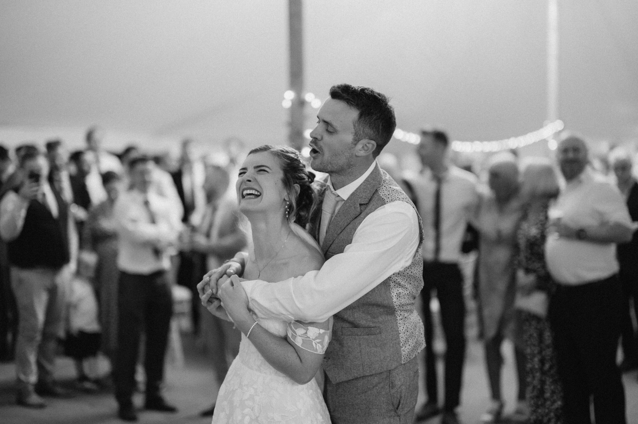 A couple joyfully embraces while singing at their wedding reception, surrounded by guests in a festively decorated venue. Capturing every heartfelt moment, their wedding photographer beautifully documents the occasion in the picturesque setting of Kent.