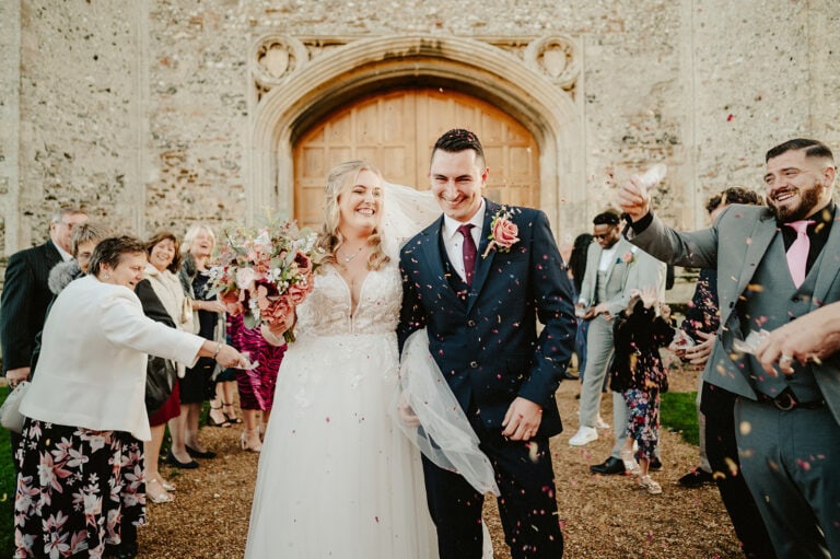 A newlywed couple smiles as they walk outside the historic stone building of Pentney Abbey, with the bride holding a bouquet and guests tossing flower petals in celebration of their joyous wedding.