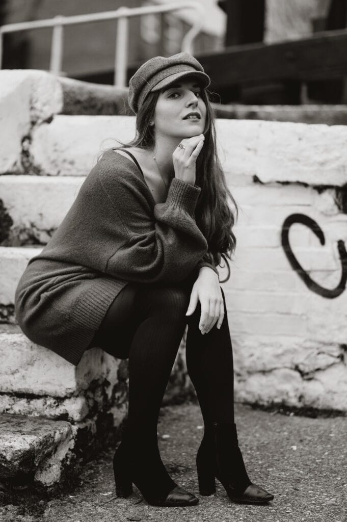 Black and white photo of a woman sitting on outdoor steps. She is wearing a hat, oversized sweater, tights, and heeled shoes, looking thoughtfully to the side with one hand near her face.