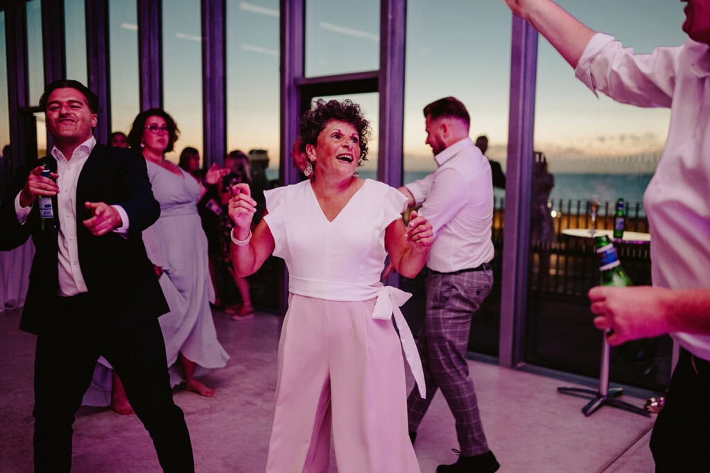A group of people dancing indoors by large windows at sunset. A woman in the center, dressed in white, is smiling and dancing, while others around her also appear to be enjoying the moment.